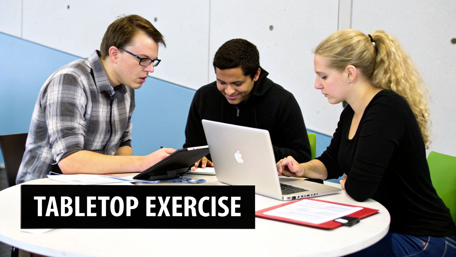 Three people collaborating at a table during a tabletop exercise with a laptop and tablet.